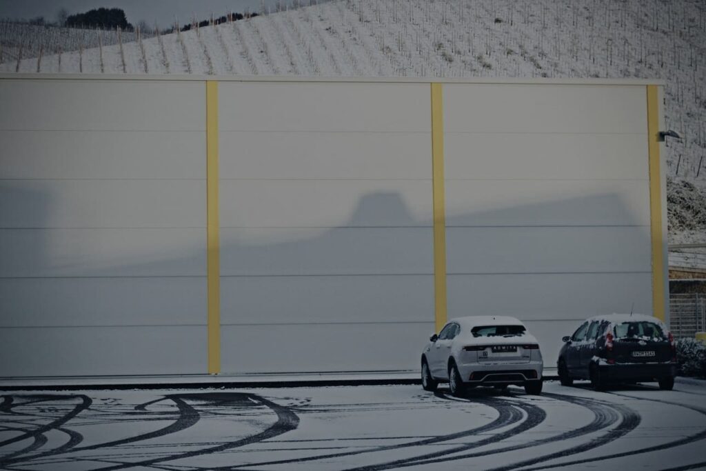 Padangų keitimo straipnio atvaizdas. Du automobiliai apsnigtoje parkavimo aikštelėje, kurioje matomos padangų paliktos vėžės sniege.